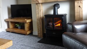 A living room with a black wood-burning stove, a wooden TV stand, a brown leather sofa, and a wooden shelf with decorative items.