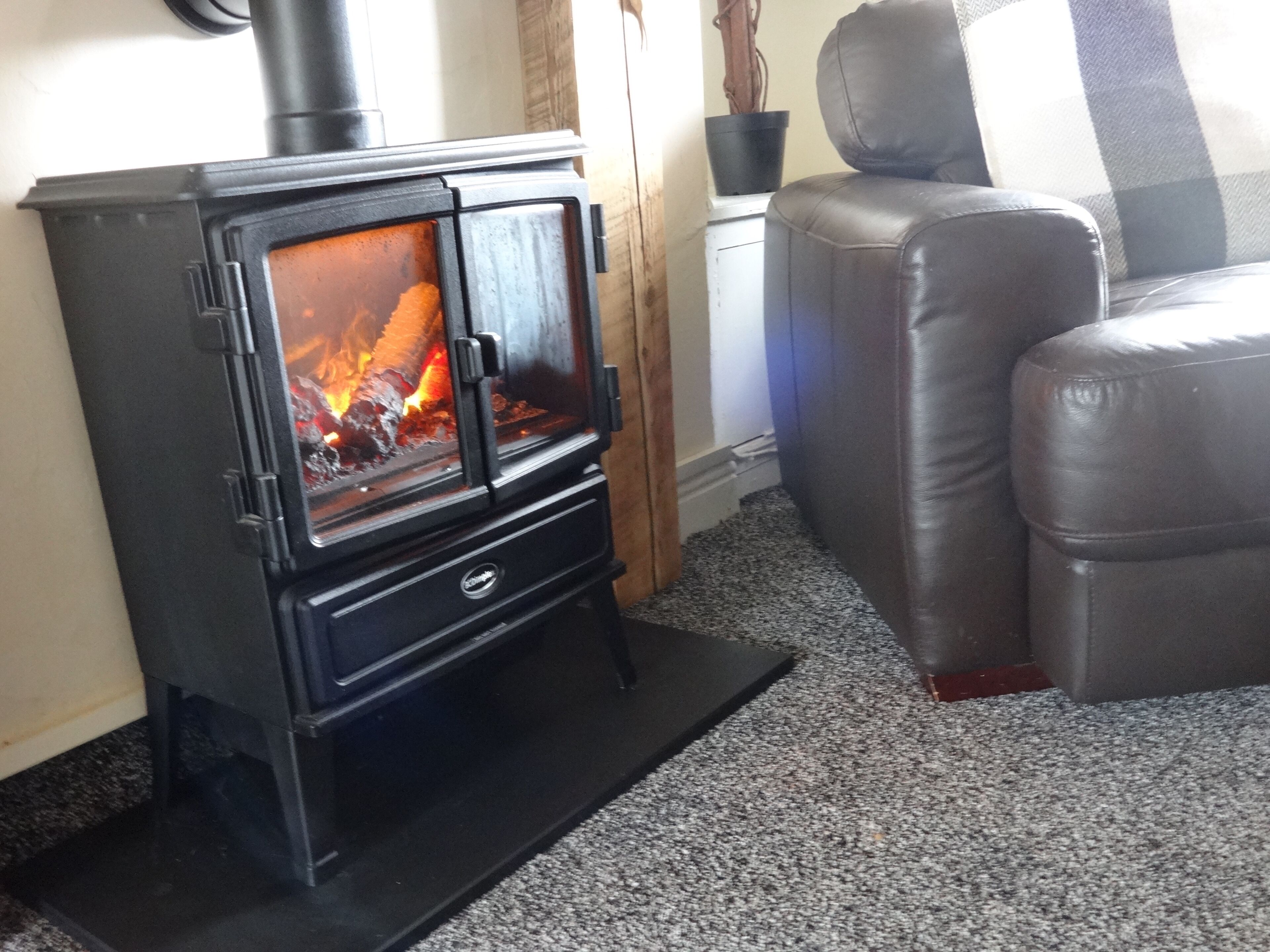 A black wood-burning stove with visible flames, a leather sofa, and a checkered cushion in a living room.