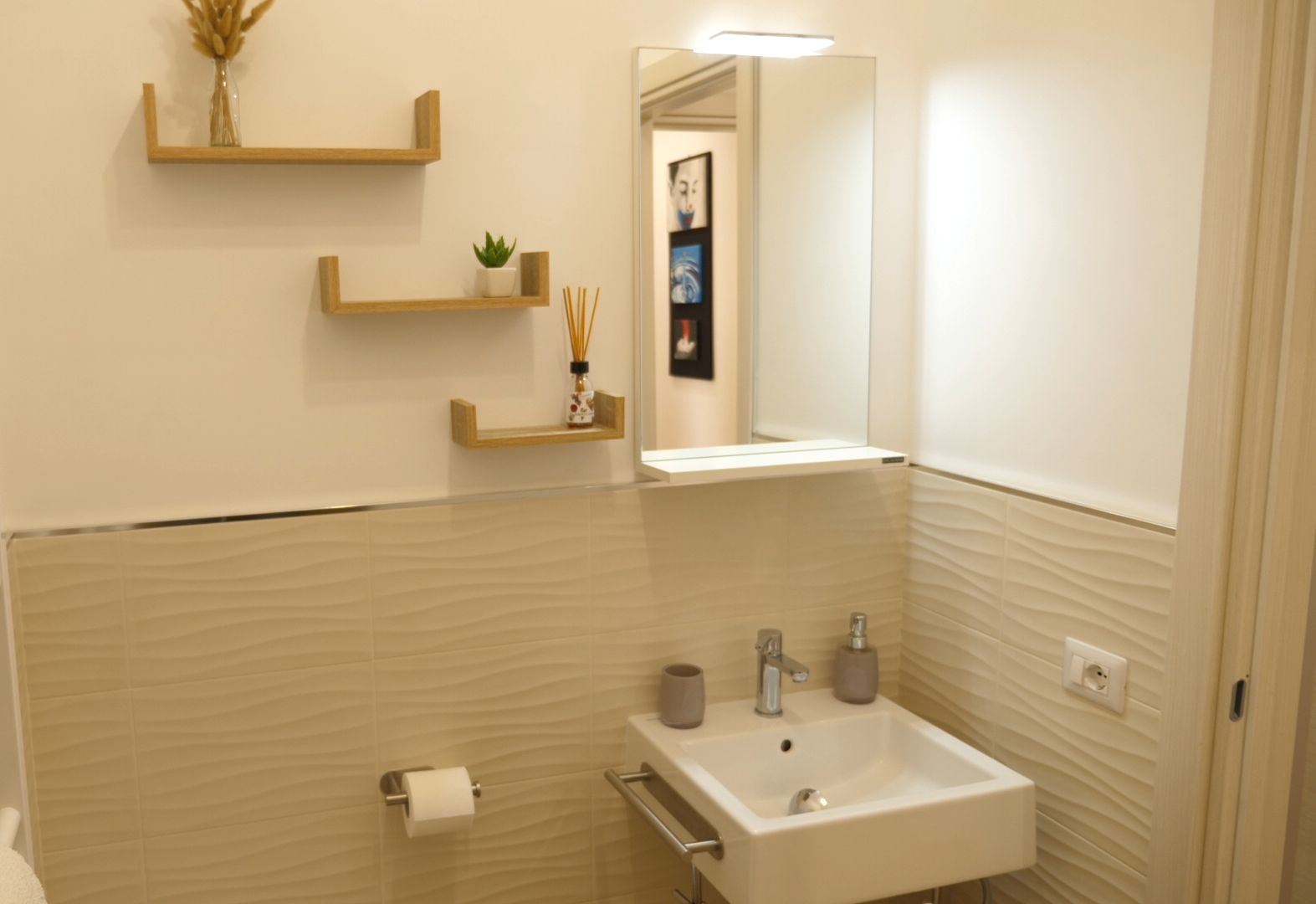 Een moderne badkamer met een wit wastafelmeubel, spiegel en handdoekhouder. Houten planken met decoratieve objecten en een toiletpapierrolhouder.