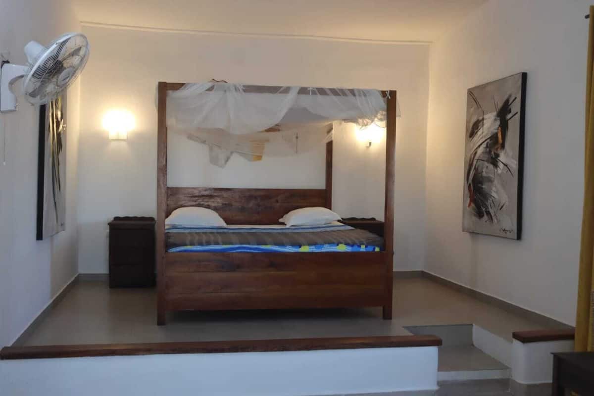 A bedroom with a four-poster bed, bedside lamps, a standing fan, and a painting on the wall.