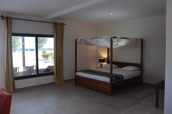 A four-poster bed with a bedside table and lamp, a window with curtains, and a view of the outdoors. - Prince Anakao  (Anakao)