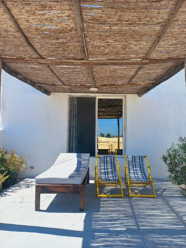 Una habitación con una cama, dos sillas a rayas, una mesita y vista a la playa por una puerta abierta.