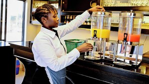 A barista preparing drinks at a coffee station with dispensers and a green basket. - Neon Hotel Ntinda (Kampala)