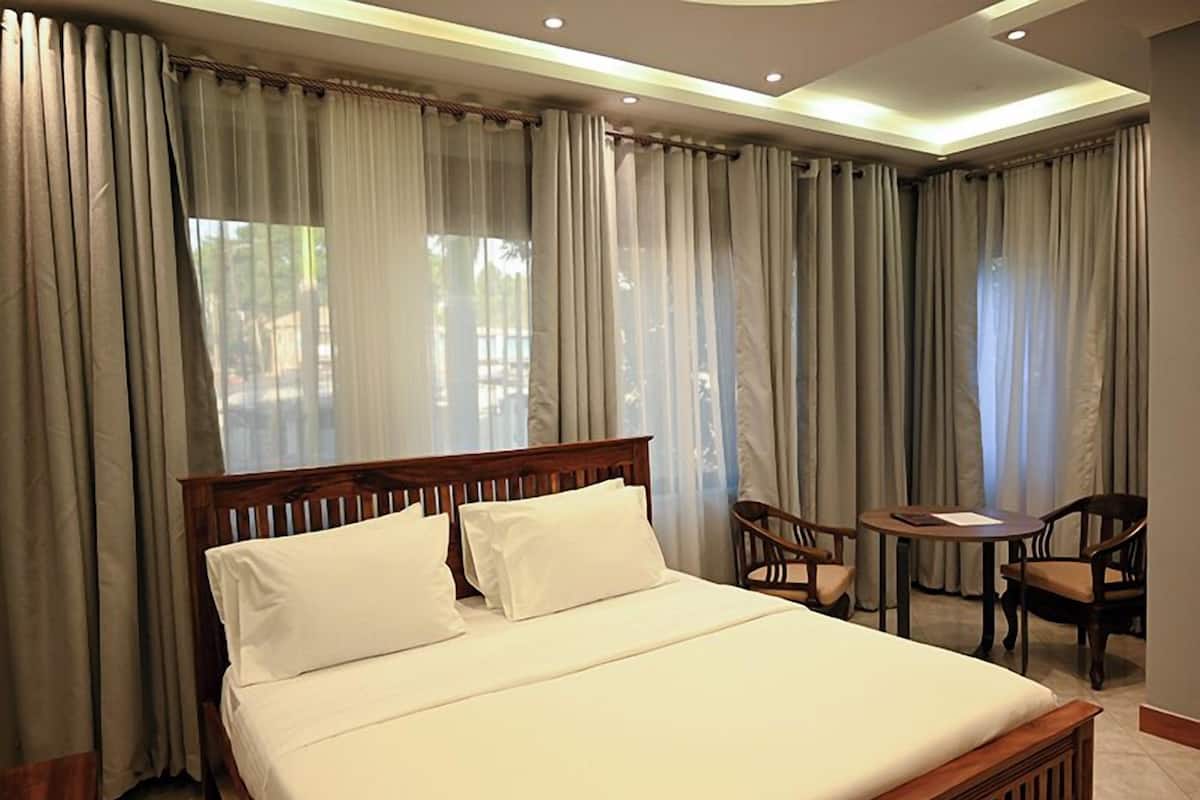 Quarto de hotel com uma cama grande, duas cadeiras, uma mesa pequena e vista para o exterior através de cortinas translúcidas.