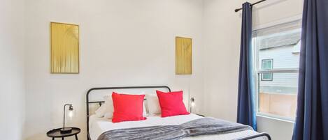 A bedroom with a bed, two red pillows, a bedside table with a lamp, and a window with curtains.