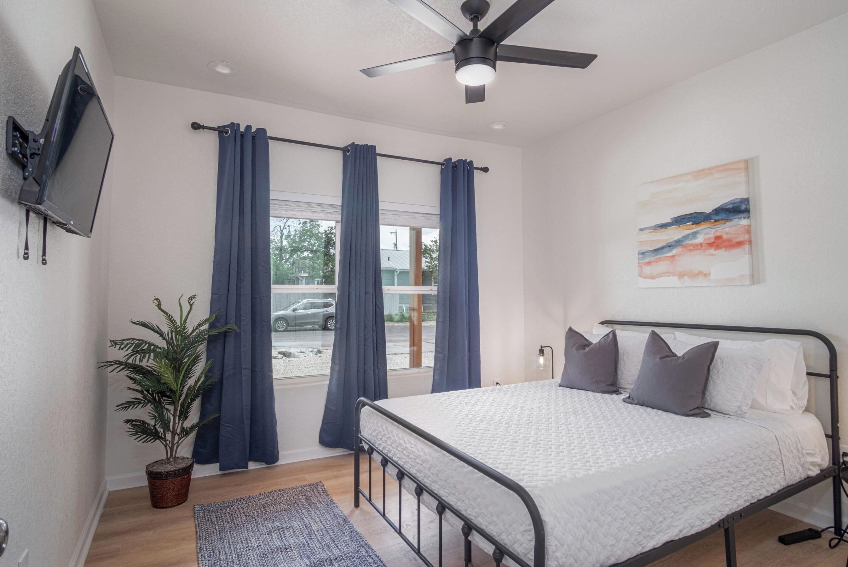 Un dormitorio con cama, ventilador de techo, televisor montado en la pared, ventana con cortinas, planta y un cuadro en la pared.
