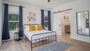 Quarto com cama, ventoinha no teto, janela com cortinas, mesa de cabeceira e porta de acesso a um banheiro.