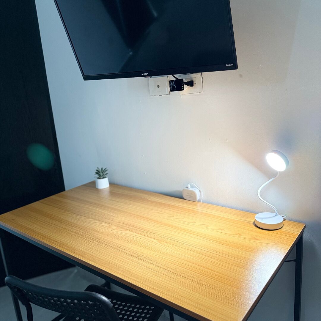 A wooden desk with a flat-screen TV mounted above it, a small potted plant, a white desk lamp, and a power outlet.