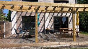 A wooden deck with outdoor furniture, a pergola, and a building with a tiled roof in the background.