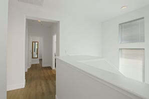 Un interior luminoso y limpio, con piso de madera, una escalera blanca y un gran ventanal con persianas.