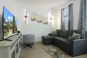 Una sala de estar moderna con un sofá seccional gris, una pequeña silla de oficina gris, un televisor de pantalla plana y obras de arte colgadas en la pared.