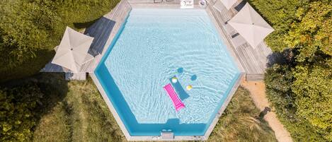 An aerial view of a hexagonal swimming pool with a wooden deck, surrounded by lush greenery.