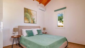Une chambre à coucher comprenant un lit, deux tables de chevet, un climatiseur fixé au mur, une fenêtre avec vue et un tableau accroché au mur.