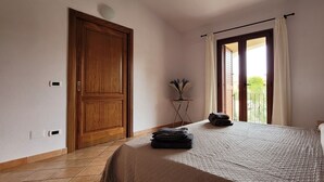 A bedroom with a wooden door, a bed with a quilt, a small table with a lamp, and a view through a window with curtains. - Nala House - Villa 800 mt from the sea (Budoni)