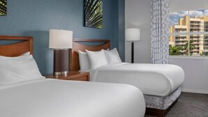 A hotel room with two beds, a nightstand with a lamp, a window with a view of buildings, and a patterned curtain.