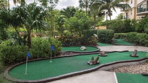 A miniature golf course with a green putting surface, surrounded by lush tropical vegetation and palm trees.