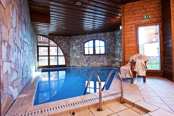 An indoor swimming pool with stone walls and wooden ceiling, a stone bench, and a door with a green exit sign.