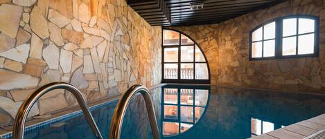 An indoor swimming pool with stone walls and a view through arched windows.