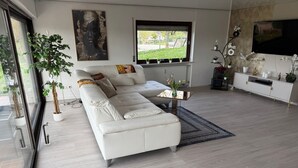 Ein modernes Wohnzimmer mit einem weißen Ecksofa, einem Couchtisch und einem großen Fenster mit Blick auf Grünflächen.