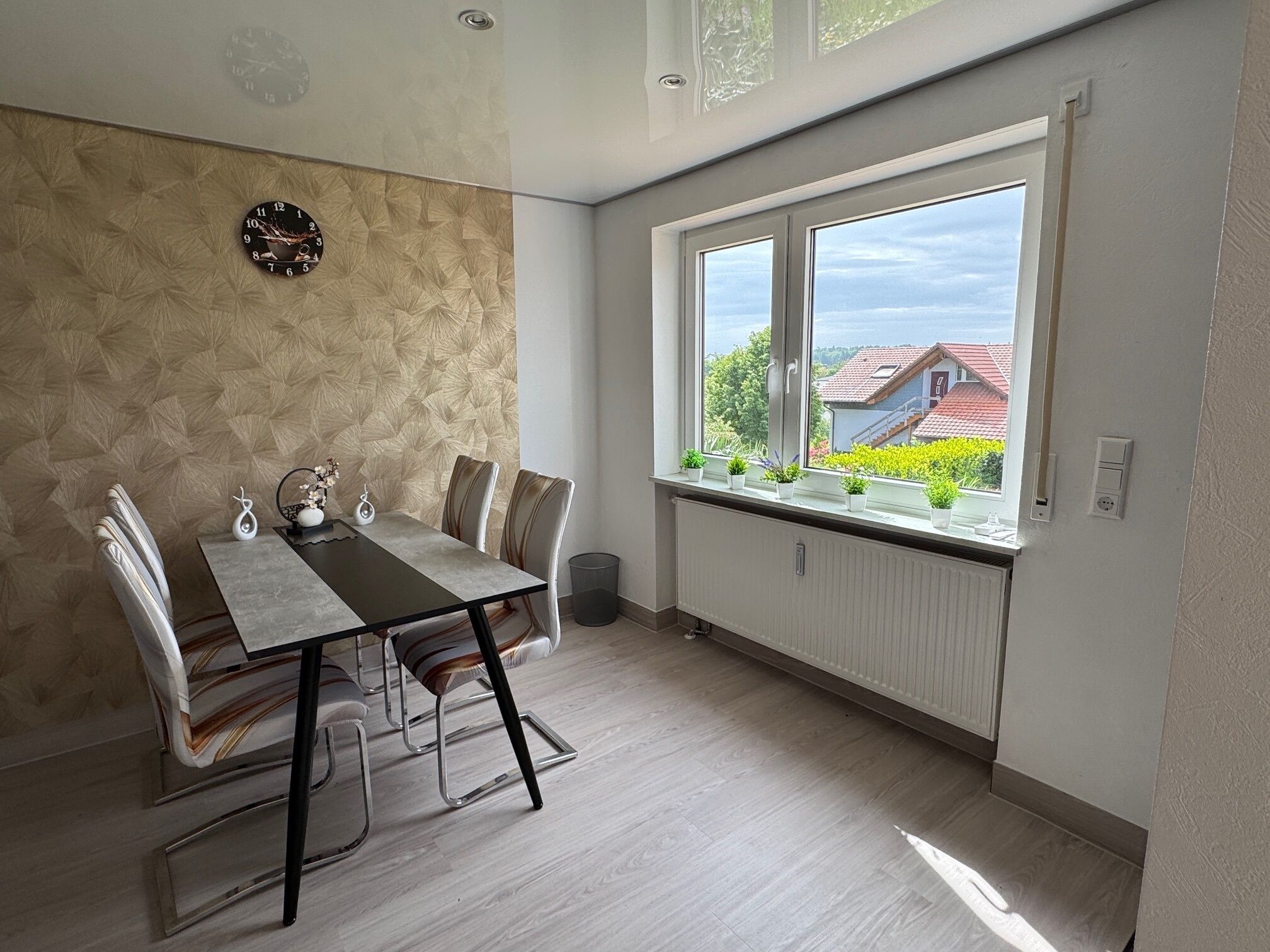 A dining area with a table, chairs, a clock, and a window with a view of buildings and greenery.