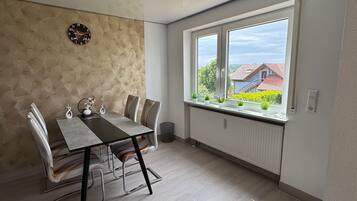 A dining area with a table, chairs, a clock, and a window with a view of buildings and greenery.