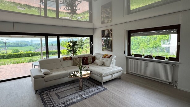 Ein großzügiges Wohnzimmer mit einem weißen Ledersofa, einem Couchtisch und Blick auf eine grüne Landschaft durch große Fenster.