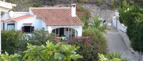 A white building with a red-tiled roof, surrounded by greenery and a stone wall.