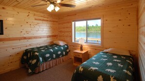 Una cabaña con dos camas, un ventilador de techo, una ventana que ofrece vistas al exterior y una pequeña mesa con una lámpara.