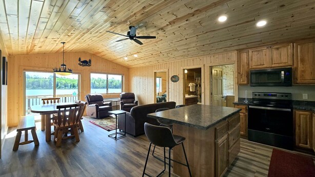 Interior de una cabaña de madera con comedor, cocina y un amplio ventanal con vistas a un lago.