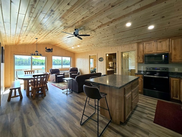 A wooden cabin interior with a dining area, kitchen, and a large window overlooking a lake.