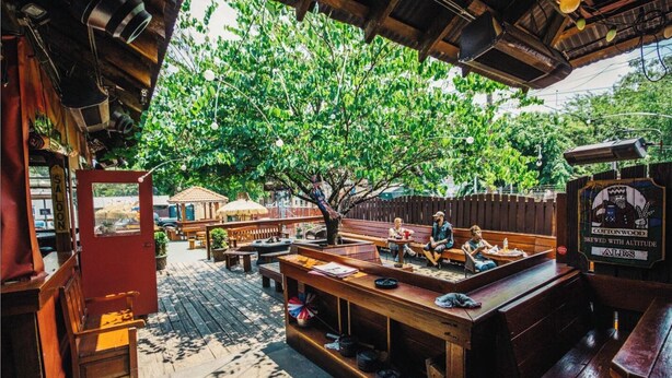 An outdoor patio with wooden tables and benches, a covered seating area, and a large tree providing shade.
