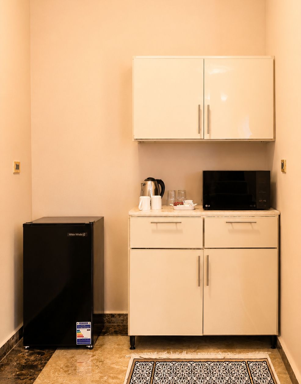 A compact kitchenette with a refrigerator, microwave, and a small countertop with a kettle and cups.