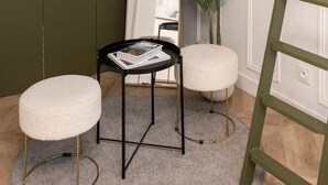 Una habitación con un tapete redondo gris, una mesita negra con revistas, dos otomanas blancas, una escalera verde y un espejo grande que refleja una planta.