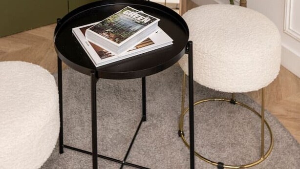 Una habitación con un espejo grande, una mesa redonda con revistas, una planta en maceta y un taburete acolchado blanco.