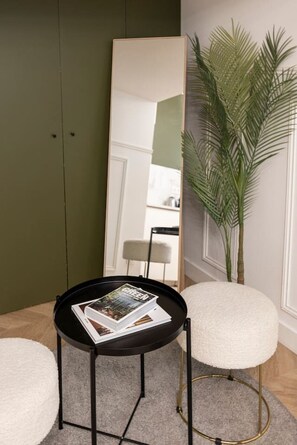 Una habitación con un espejo grande, una mesa redonda con revistas, una planta en maceta y un taburete acolchado blanco.