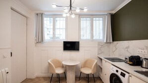 Una cocina compacta con lavadora, una pequeña mesa de comedor con sillas, un televisor montado en la pared y un ventanal con cortinas.