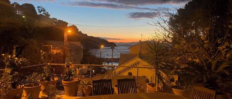 Une terrasse avec une table ronde et des chaises, entourée de plantes en pot et offrant une vue sur la côte et les couchers de soleil.