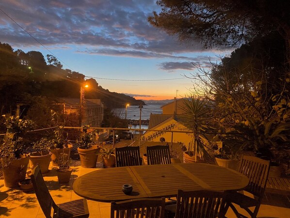 A patio with a round table and chairs, surrounded by potted plants and overlooking a coastal area with a sunset view.