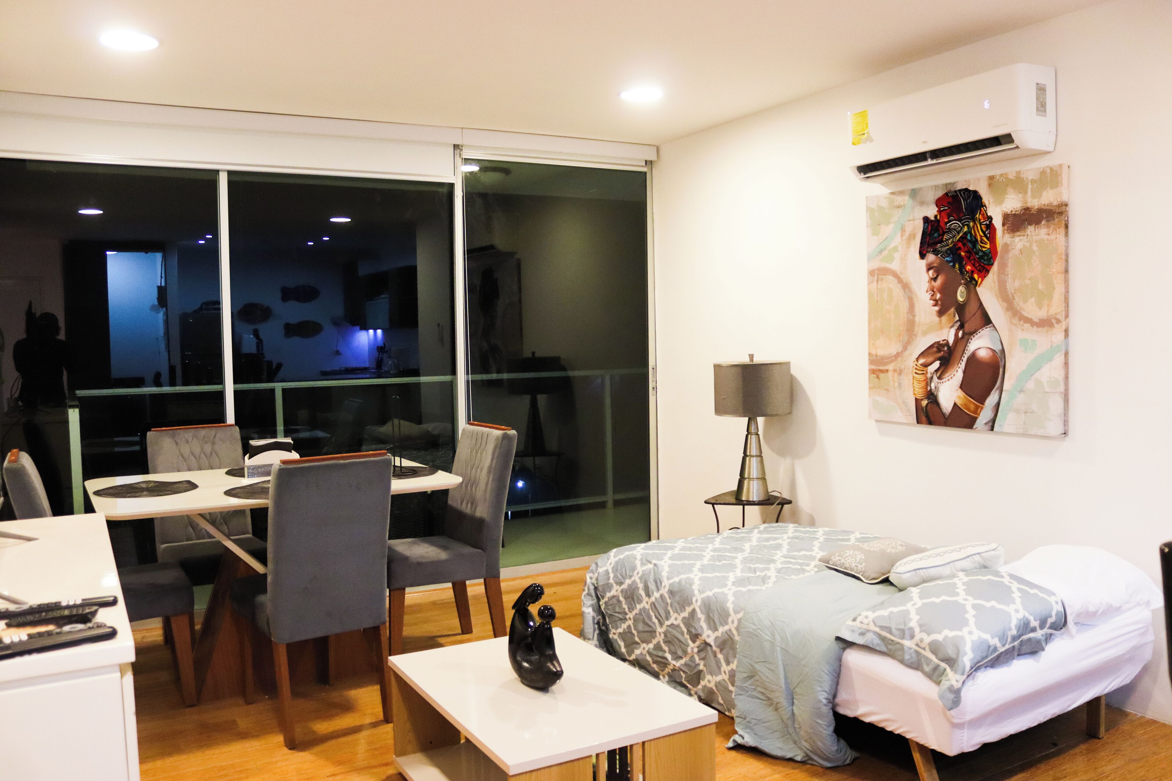 A modern hotel room with a dining area, a bed with patterned bedding, a desk with a lamp, and a large window with a view.