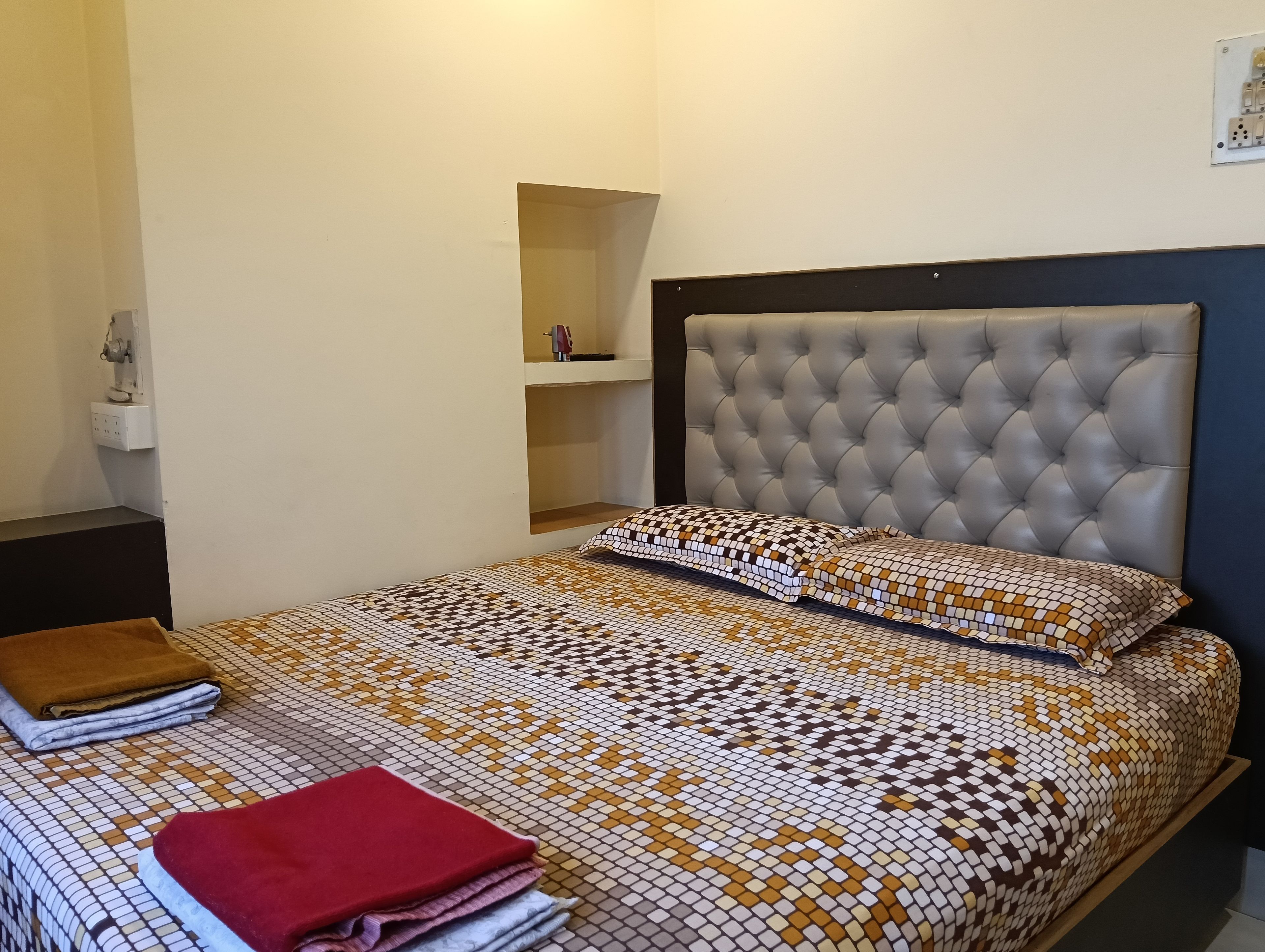 A bedroom with a bed, a nightstand, a wall socket, and a shelf with a red object.