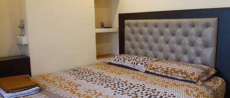 A bedroom with a bed, a nightstand, a wall socket, and a shelf with a red object.