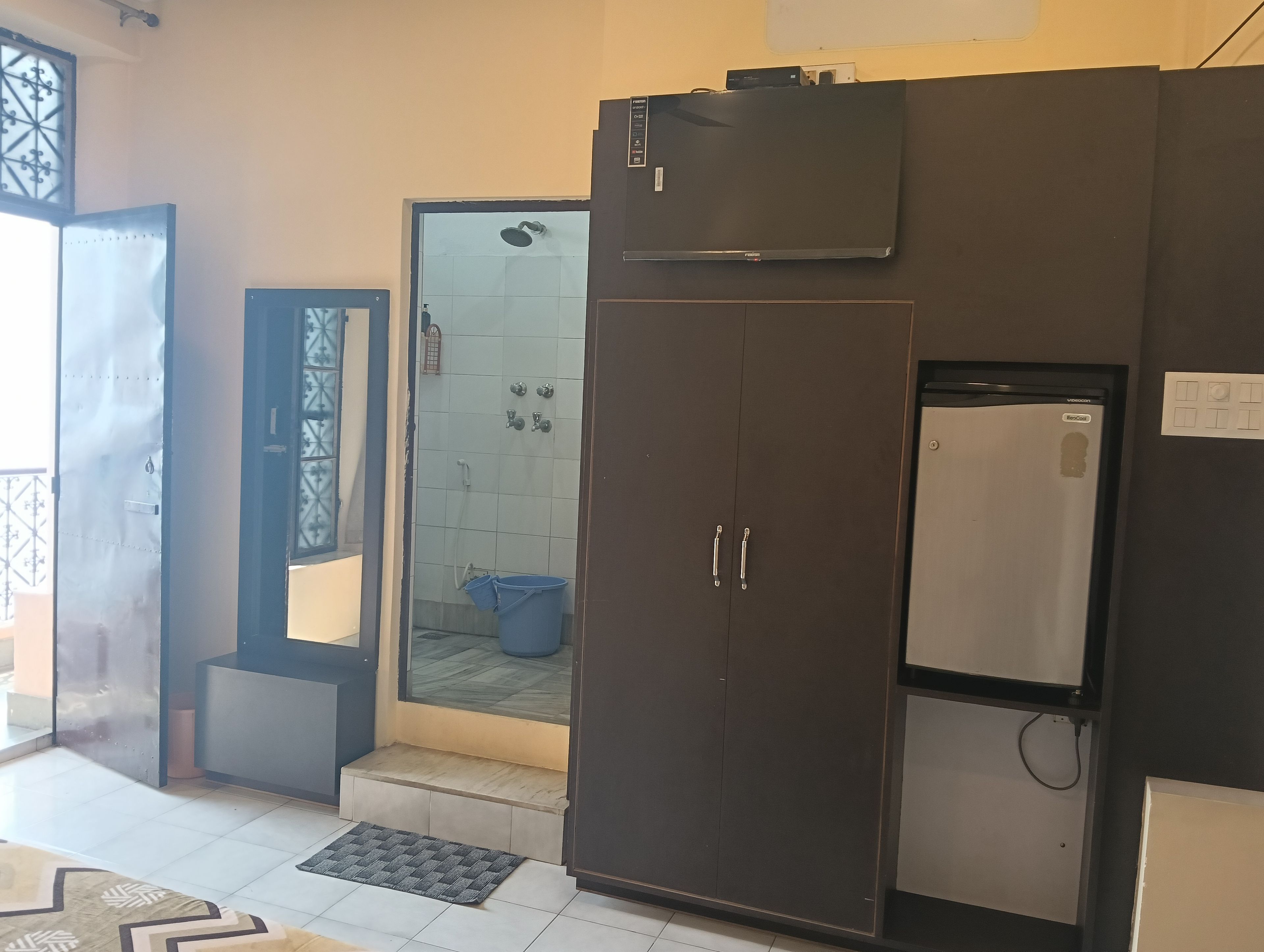 Habitación con un armario grande, un refrigerador pequeño, una puerta con ventanal y una zona de ducha con azulejos.