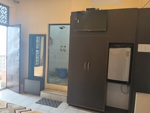 A room with a large wardrobe, a small refrigerator, a door with a window, and a tiled shower area. - Scindhia Guest House (Varanasi)