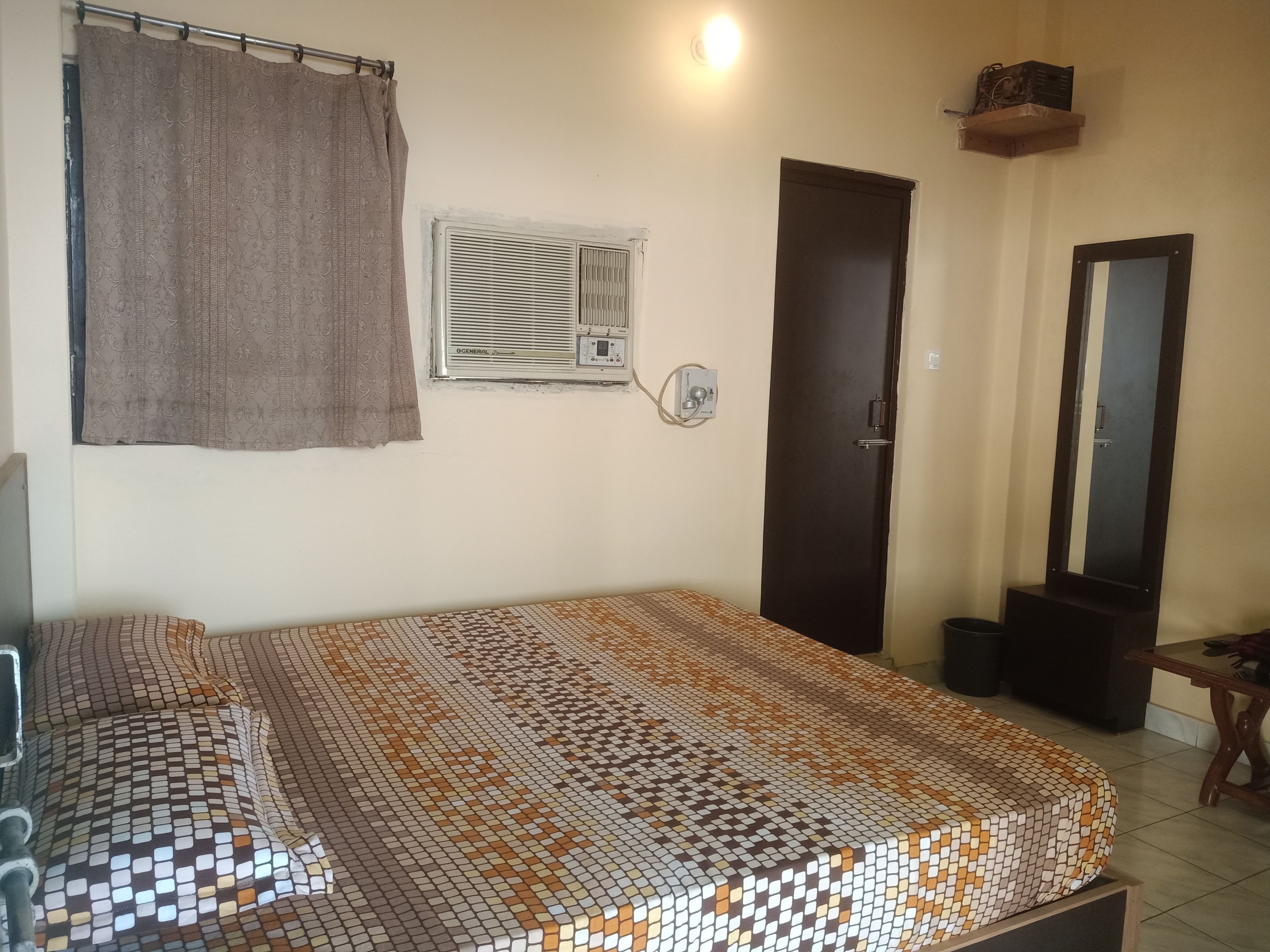 A room with a bed, an air conditioning unit, a door, a mirror, and a small table.