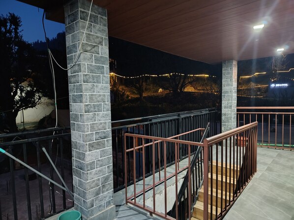 A covered outdoor area with a stone pillar, a metal railing, and a staircase. - Bodh Niwas - Unit of Nest Community (Manali)