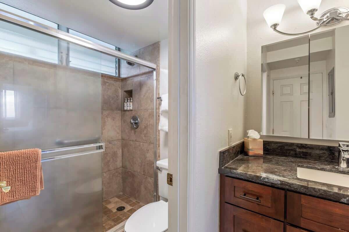 A bathroom with a glass shower enclosure, a toilet, a sink with a dark countertop, and a mirror with lights.