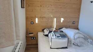 Una habitación pequeña y acogedora con techo de madera, una cama con almohadas y un bulto encima, y un estante con un par de zapatos.