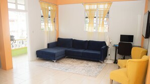 A living room with a blue sectional sofa, a yellow armchair, a flat-screen TV, and a small table. - Elite Island Breeze 2-bed apt in Jimmit, WiFi, AC, Free Parking,Central location (Jimmit)