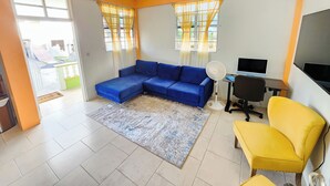 A living room with a blue sectional sofa, a yellow armchair, a small desk with a computer, and a TV. - Elite Island Breeze 2-bed apt in Jimmit, WiFi, AC, Free Parking,Central location (Jimmit)