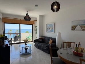A living room with a sofa, a coffee table, a dining table, and a view of the sea through a sliding door.